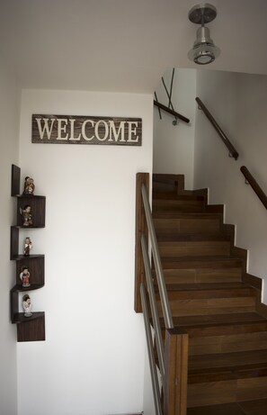 Una escalera de madera con barandilla de metal que conduce a una plataforma con un cartel de 'WELCOME' y pequeños objetos decorativos en estantes.