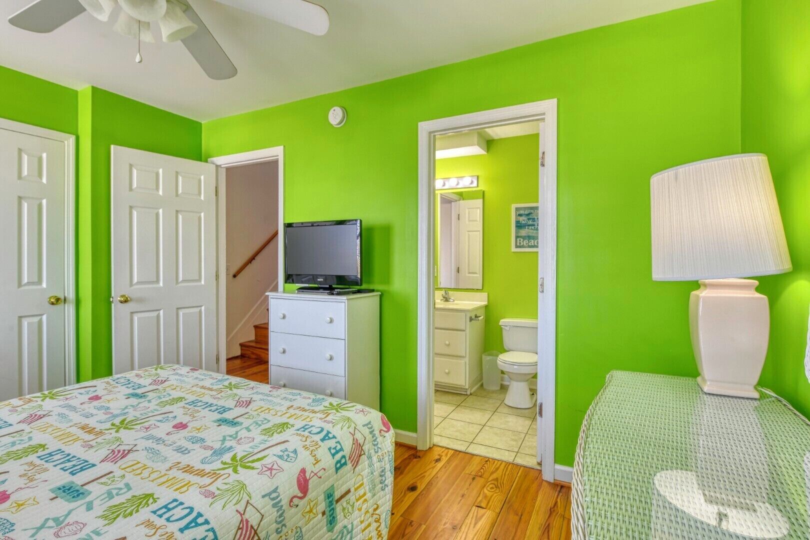 A room with a bed, a dresser, a television, a bathroom with a toilet and sink, and a ceiling fan.