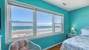 A room with a large window overlooking a beach, a bed with a patterned bedspread, a chair, and a nightstand with a lamp. - Against The Wind Side B, Oceanfront Home, Awesome view of Garden City Pier (Garden City Beach)