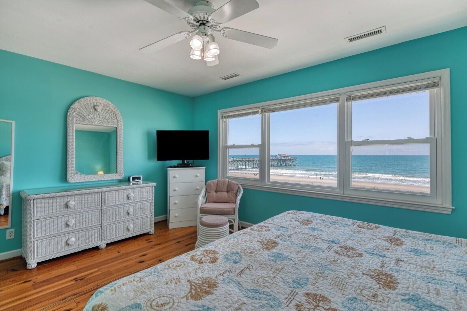 A bedroom with a bed, dresser, chair, TV, and a large window with a beach view.