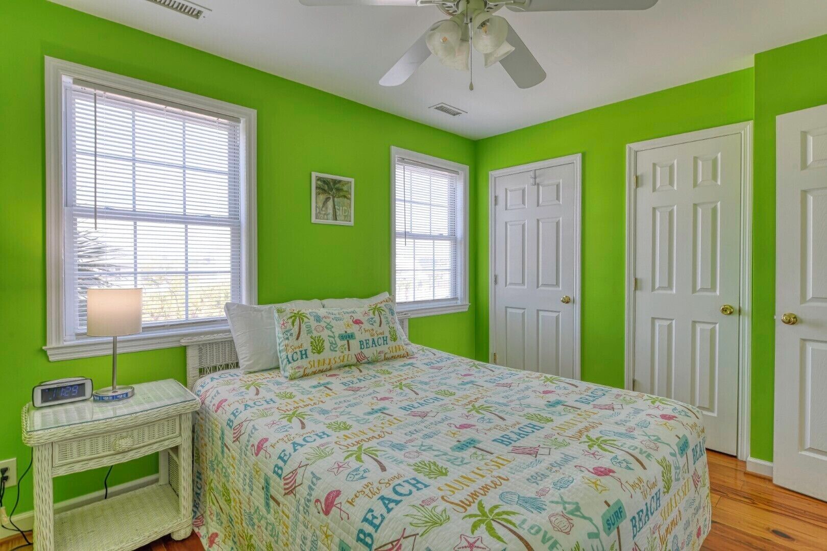 A bedroom with a bed, two windows, a door, a bedside table with a lamp, and a wall clock.