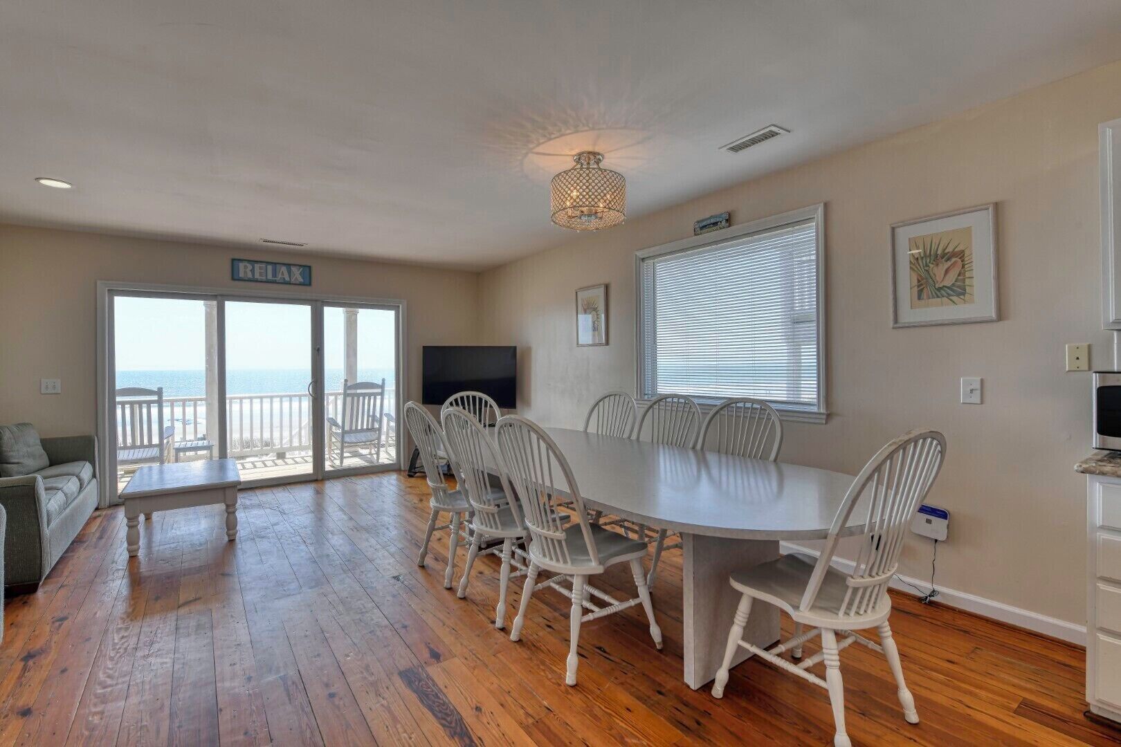A spacious living area with a large dining table, white chairs, a flat-screen TV, and a balcony with a view of the ocean.