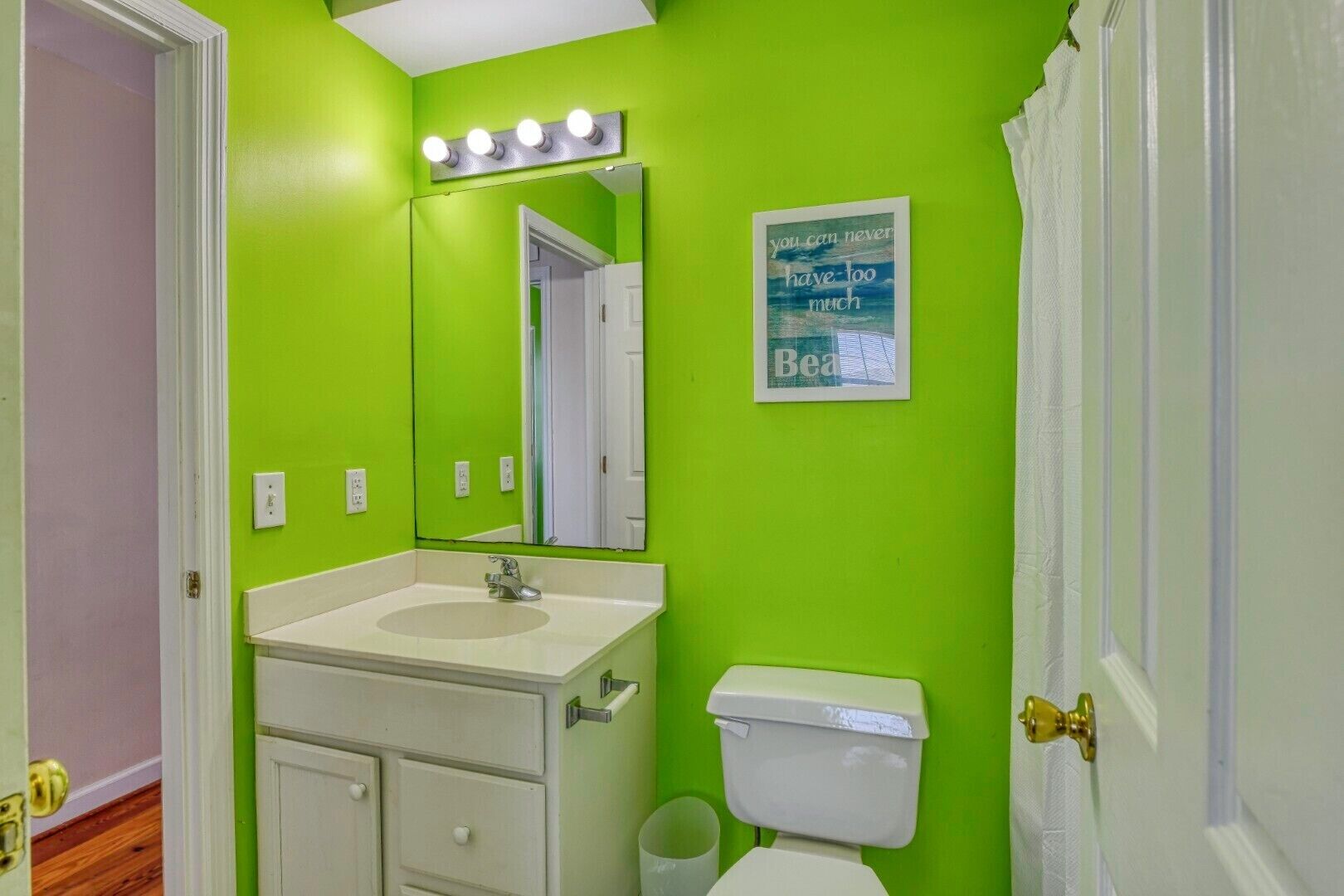 A bathroom with a green wall, a white sink, a mirror with lights, a toilet, and a poster.