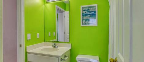 A bathroom with a green wall, a white sink, a mirror with lights, a toilet, and a poster.