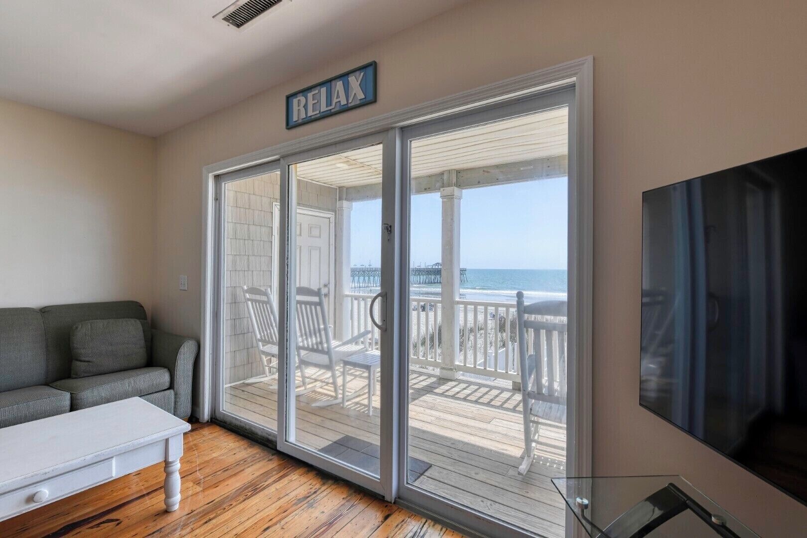 A room with a sliding glass door leading to a balcony with beach views, a grey sofa, and a white table.