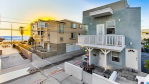 A coastal residential area with a modern two-story building featuring a balcony, a patio with outdoor seating, and a view of the ocean.