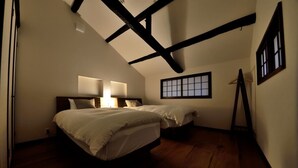 A bedroom with two beds, wooden flooring, a sloped ceiling with exposed beams, a window with a grid pattern, and a small table with a chair. - Machiya Kamo 7 (Kyoto)
