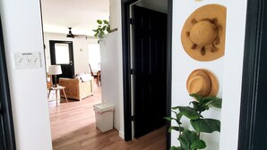 Un couloir avec un plancher en bois, une porte noire et un porte-chapeaux fixé au mur, sur lequel sont posés trois chapeaux. Un pot de fleurs se trouve à droite et une petite poubelle à gauche.