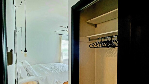 Une chambre à coucher avec un lit, un tabouret en bois, une lampe suspendue et une armoire avec un portant à vêtements.