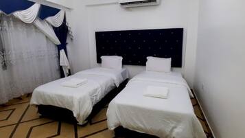 Habitación con dos camas individuales, ropa de cama blanca, cabecero oscuro, unidad de aire acondicionado y suelo estampado.