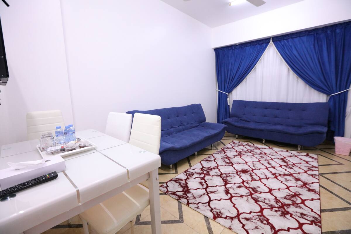 A hotel room with a white bed, two blue sofas, a white table with a tray of items, and a patterned rug.