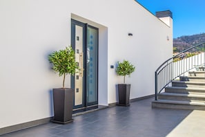 Enter A&M HOUSE through a sleek glass door, flanked by lush greenery and a staircase leading to a modern interior.