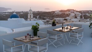 A rooftop terrace with a table set for dining, overlooking a coastal town with white buildings and a church with a blue dome. - Bliss Mansion with Swimming Pool, Cave Hot Tube & Sunset view  (Pyrgos Kallistis)