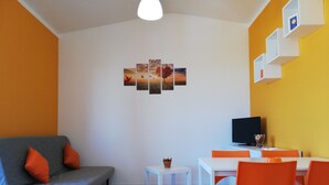A modern living room with a grey sofa, orange cushions, a white coffee table, and orange chairs. A flat-screen TV is mounted on the wall, and there are four framed pictures above the sofa. - Between Tropea and Pizzo: "La Gramigna Villette" in Calabria, 600 meters from the sea (Briatico)