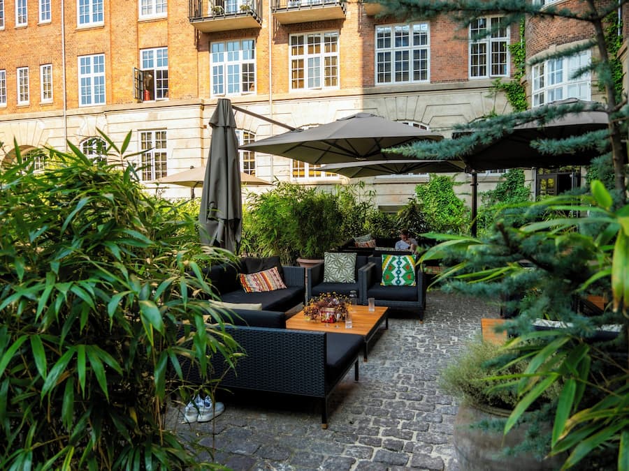 An outdoor seating area with sofas, a table, and umbrellas, surrounded by greenery and a brick building in the background.