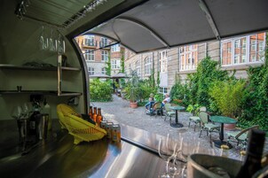 Vista desde el interior de un vehículo a un patio con mesas y sillas, un edificio con grandes ventanales y una zona cubierta con sombrillas.