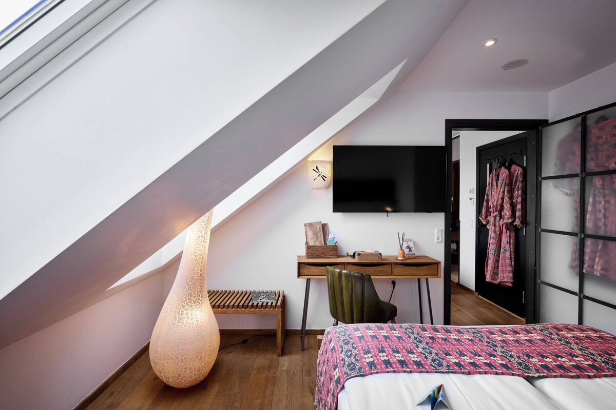 A modern bedroom with a sloped ceiling, a large bed, a desk, a chair, a wall-mounted TV, and a wardrobe.