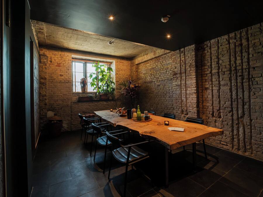 Un comedor con una mesa de madera larga, sillas negras, pared de ladrillo y ventana con plantas.
