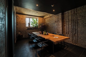 Un comedor con una mesa de madera larga, sillas negras, pared de ladrillo y ventana con plantas.