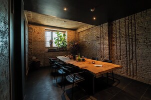 Un comedor con una mesa de madera larga, sillas negras, pared de ladrillo y ventana con plantas.