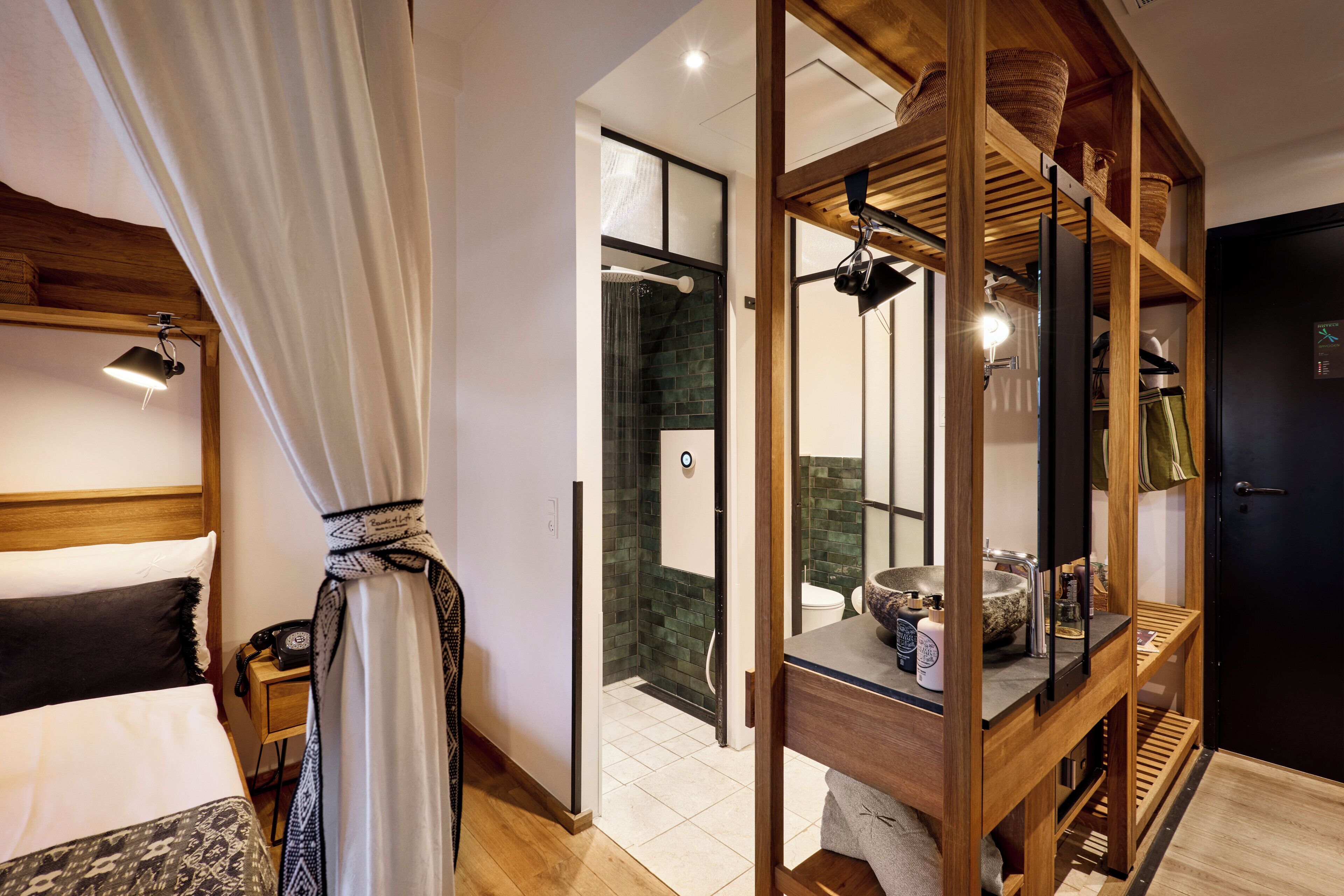 A modern hotel room with a bed, a wooden headboard, a shower area, and a wooden shelf with baskets.