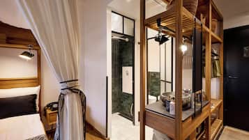A modern hotel room with a bed, a wooden headboard, a shower area, and a wooden shelf with baskets.