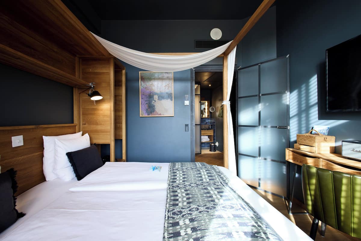 A modern hotel room with a large bed, a wooden headboard, a desk with a Marshall speaker, and a TV mounted on the wall.