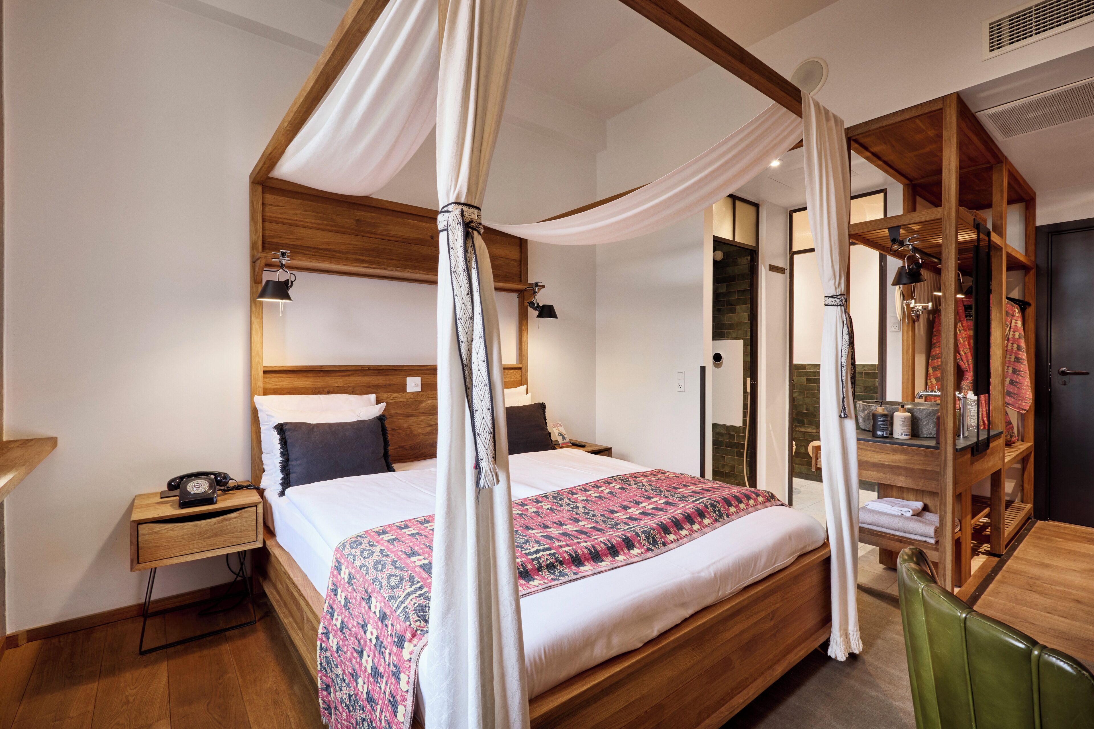 Una cama con dosel, una mesita de noche de madera con teléfono, una silla verde y un armario de madera con espejo.