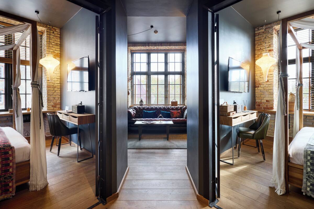 Une chambre d’hôtel moderne avec un plancher en bois, un lit recouvert d’une courtepointe à motifs, un bureau avec une chaise, un canapé avec des coussins et une grande fenêtre avec des stores.