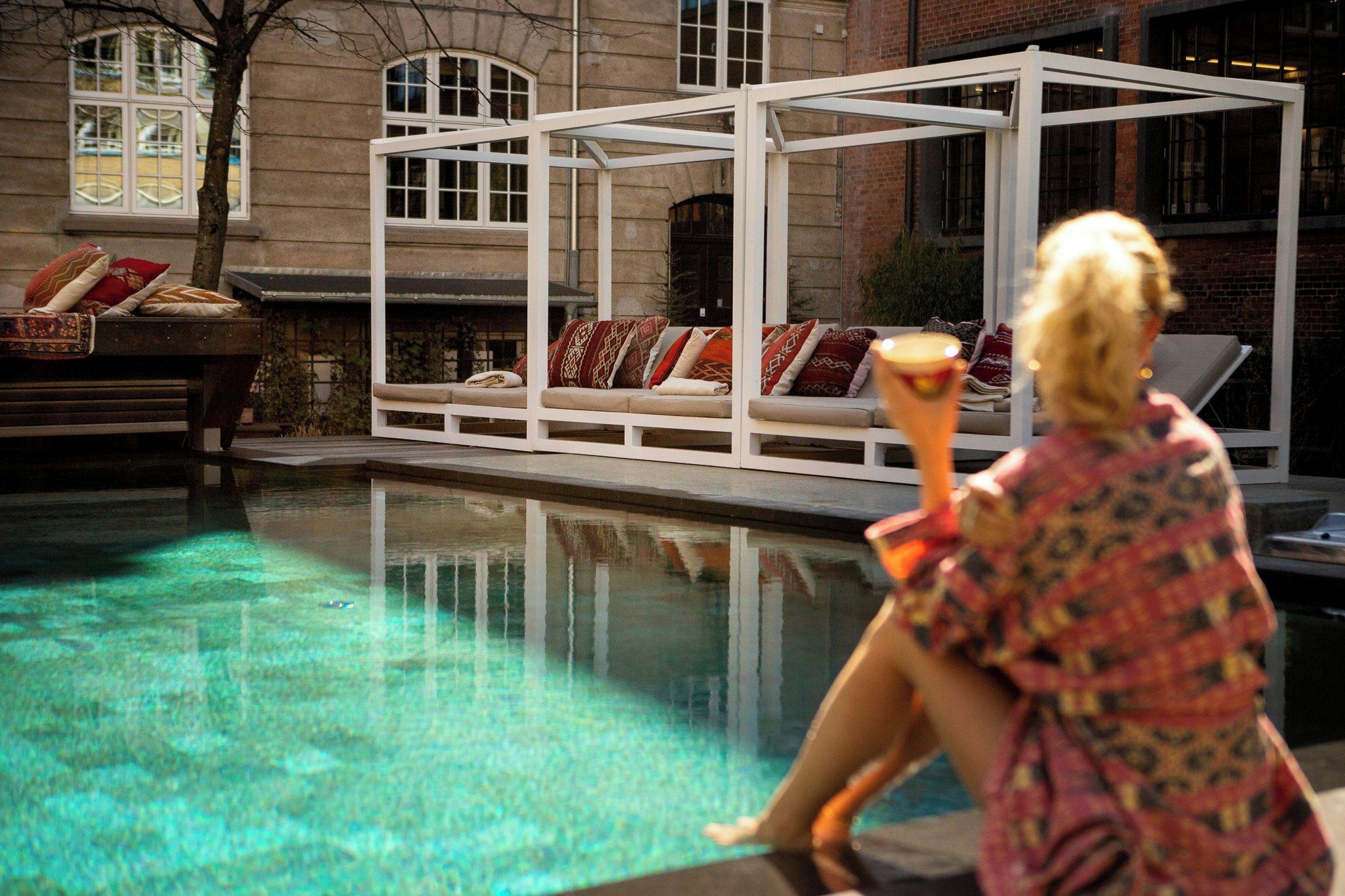 A pool area with a white structure, a lounge chair, and a person holding a drink.