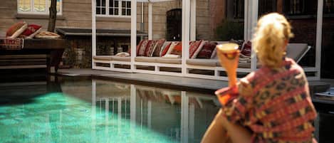 A pool area with a white structure, a lounge chair, and a person holding a drink.