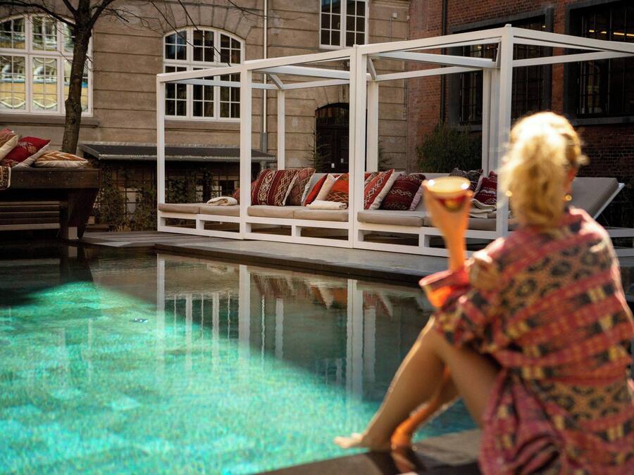 Zona de piscina con una estructura blanca, una silla de descanso y una persona sosteniendo una bebida.