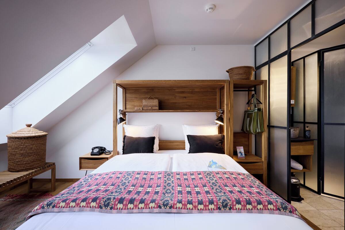 A modern bedroom with a sloped ceiling, a wooden headboard, a large bed with patterned bedding, a bedside table with a lamp, and a small shelf with a basket.