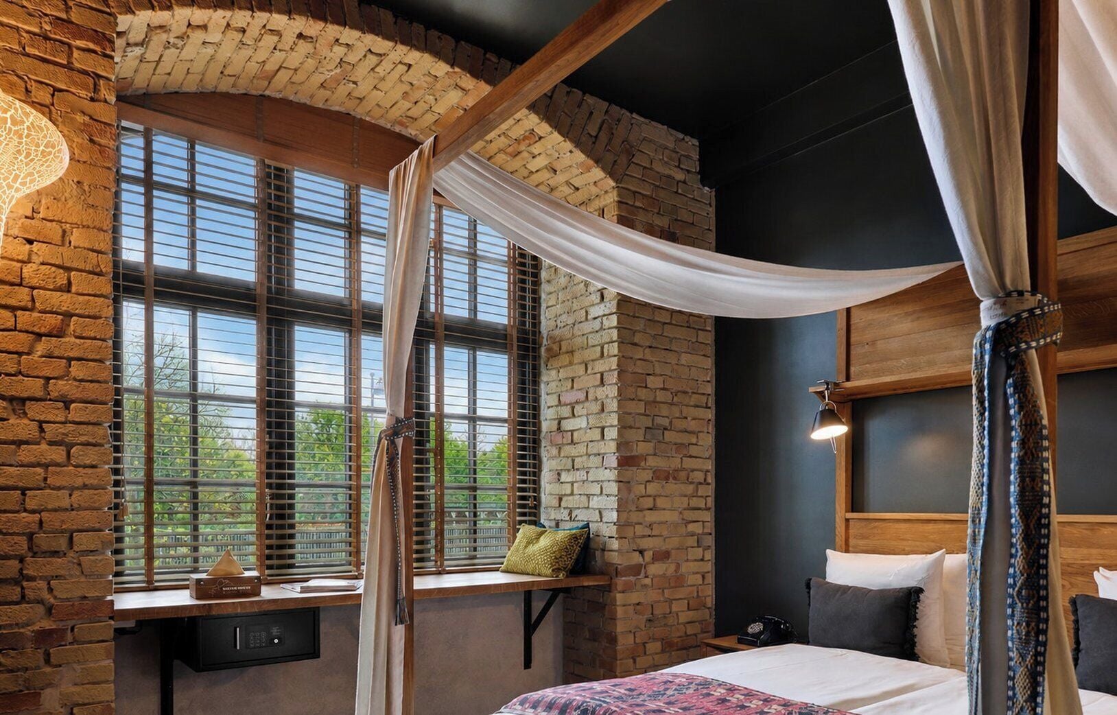 A bedroom with a brick wall, a large window with blinds, a bed with a white bedspread, and a bedside table with a lamp.