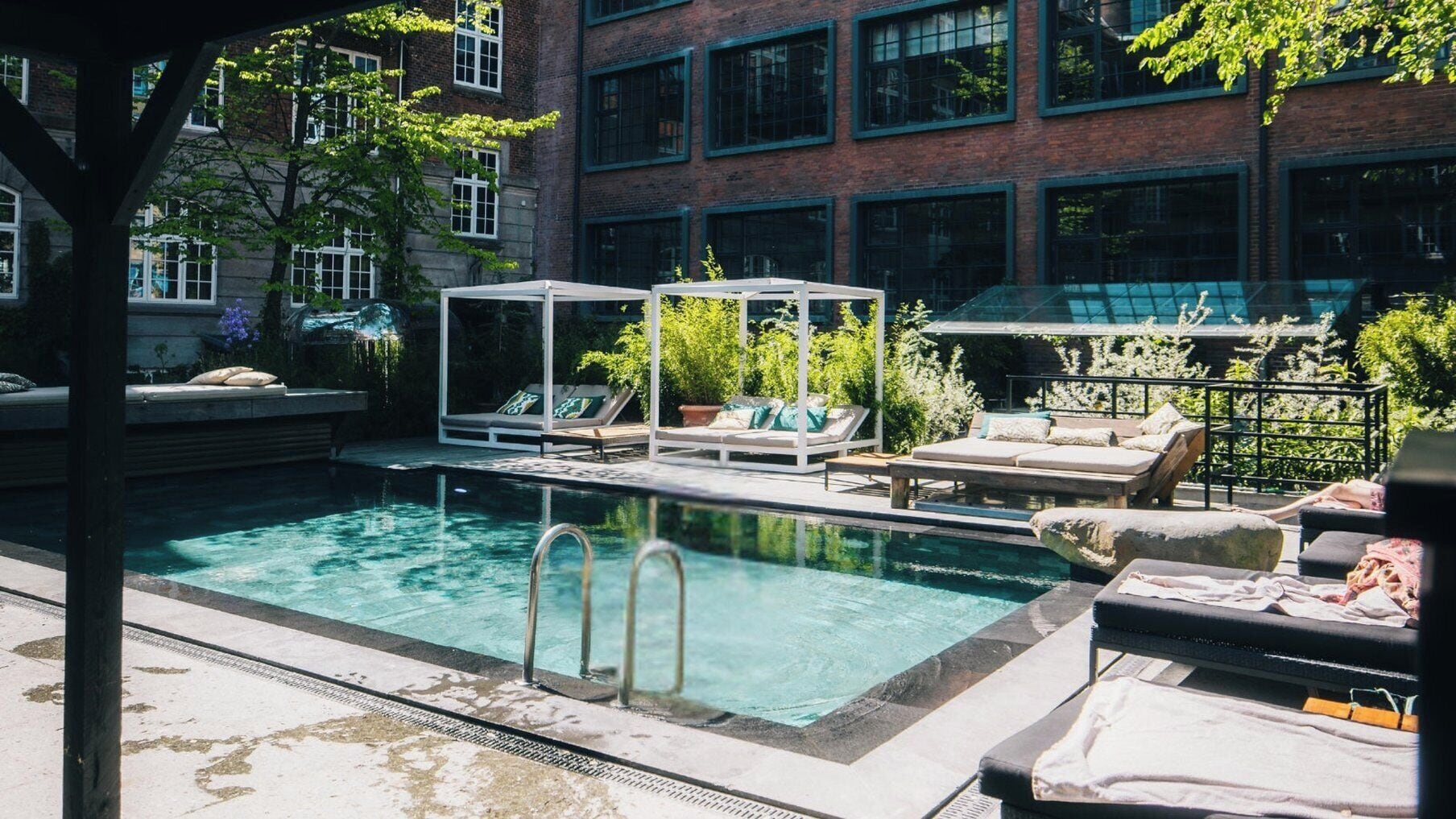 A rooftop pool with lounge chairs and a covered seating area, surrounded by greenery and a brick building.