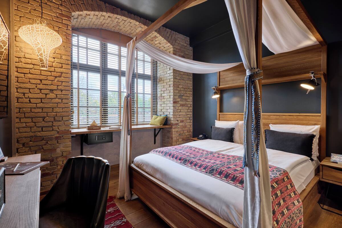 A bedroom with a brick wall, a large bed with a canopy, a desk with a chair, and a window with blinds.
