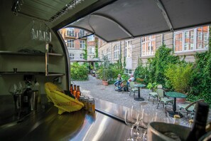 Blick aus einem Fahrzeug auf einen Innenhof mit Tischen und Stühlen, ein Gebäude mit großen Fenstern und ein überdachter Bereich mit Sonnenschirmen.