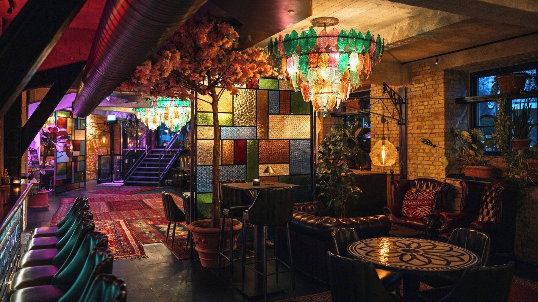 A cozy lounge area with a staircase, a chandelier, and a variety of seating options including a round table and cushioned chairs.