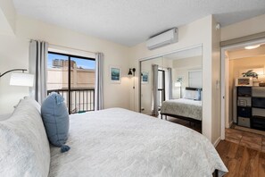 A bedroom with a large bed, a window with curtains, an air conditioning unit, a mirror, and a view of the outside.