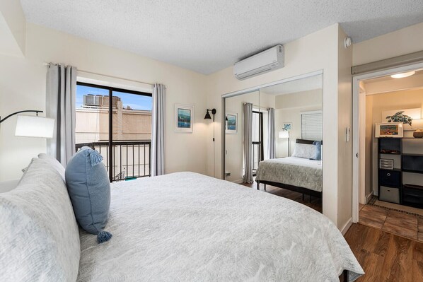 A bedroom with a large bed, a window with curtains, an air conditioning unit, a mirror, and a view of the outside.
