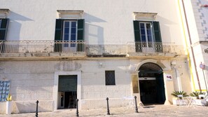 Un edificio con fachada de piedra, dos ventanas con contraventanas verdes, un balcón con barandilla metálica y una puerta con un cartel que dice "Restaurant La Piazza".