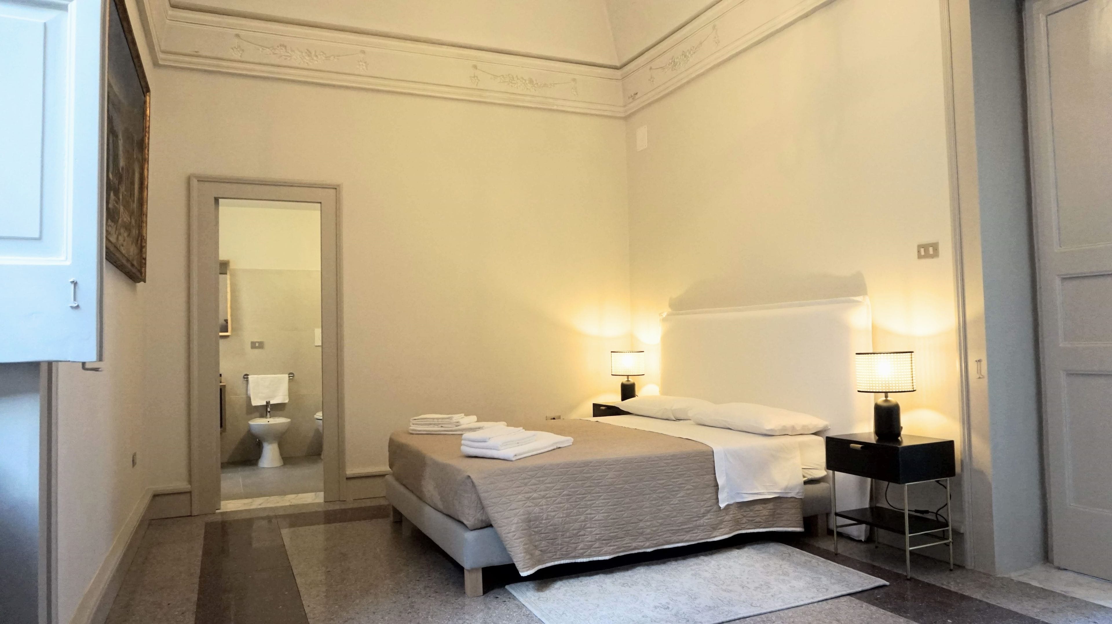 Ein Hotelzimmer mit Bett, zwei Nachttischlampen, einem Nachttisch, einem Badezimmer mit Toilette und Waschbecken sowie einer Tür.