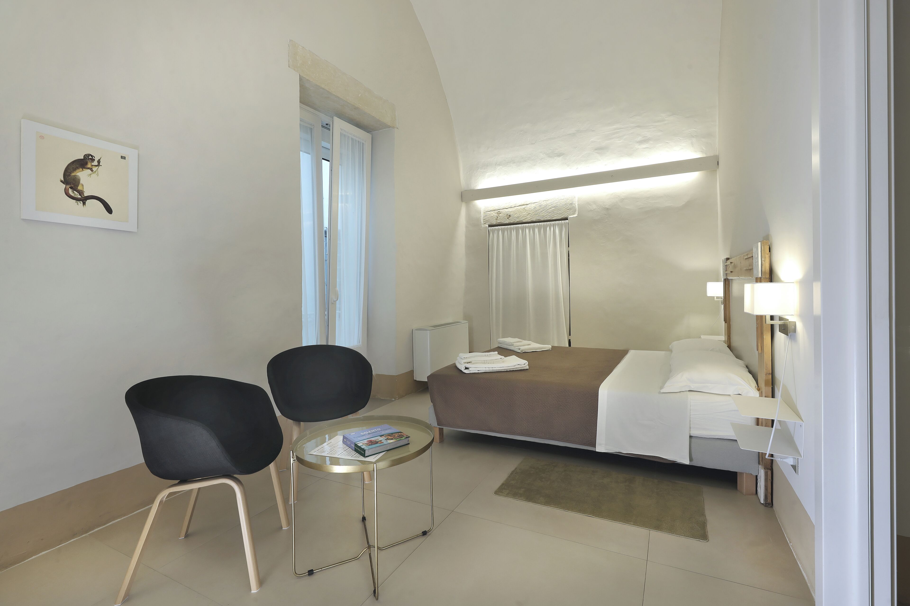 A hotel room with a bed, two chairs, a small table, a bedside lamp, and a window with curtains.