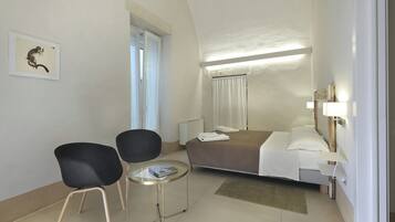 A hotel room with a bed, two chairs, a small table, a bedside lamp, and a window with curtains.