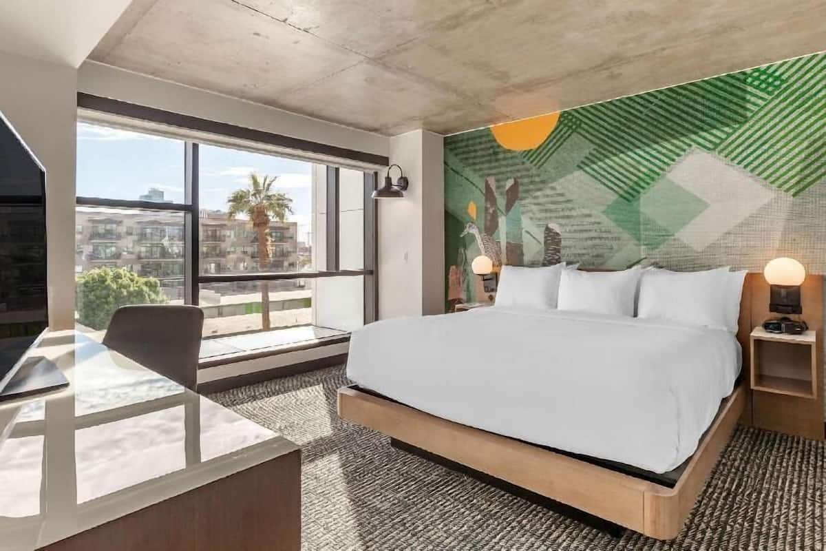 A modern hotel room with a large bed, a desk, and a view of the city through large windows.