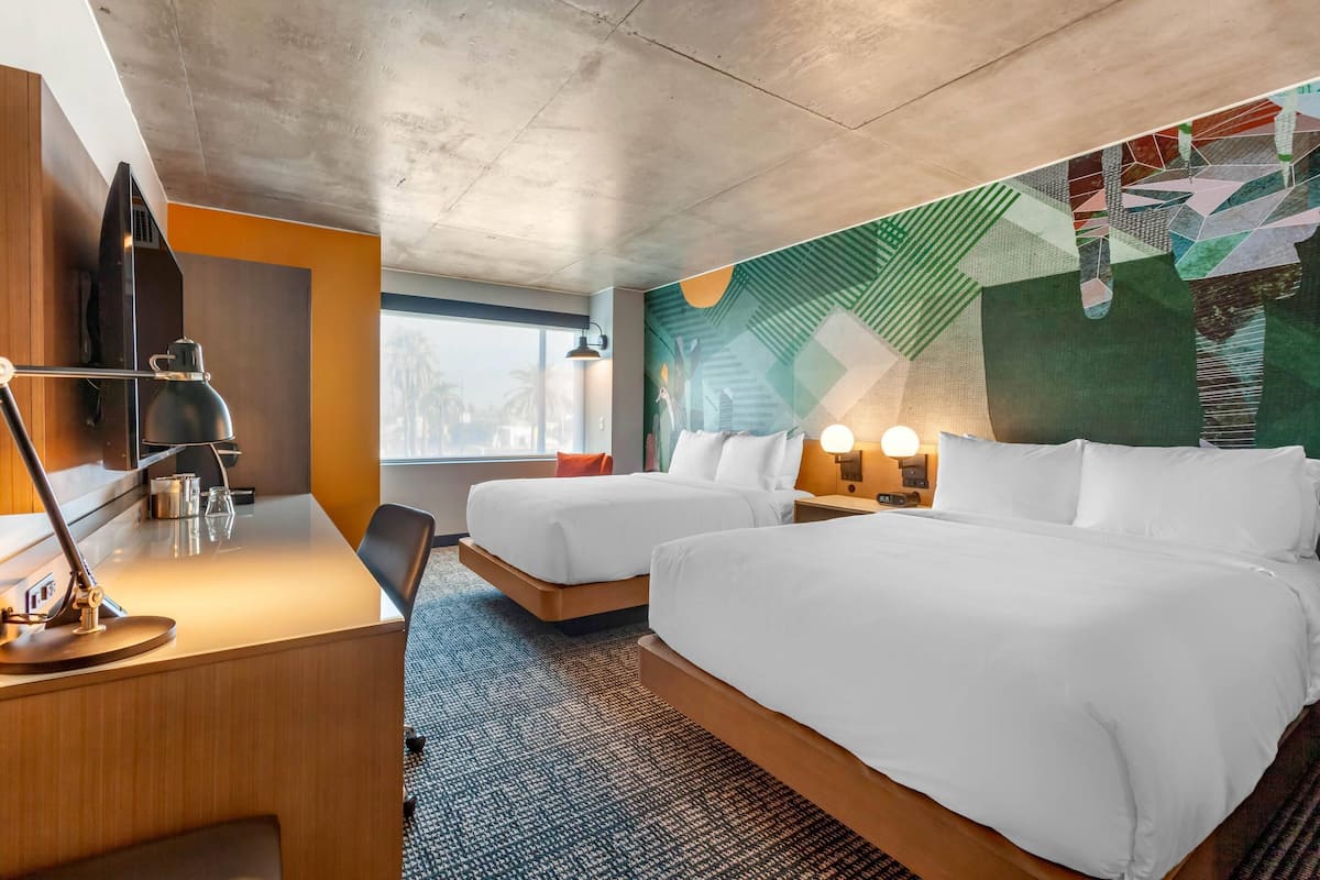 A hotel room with two beds, a desk with a lamp, a TV, and a wall mural.
