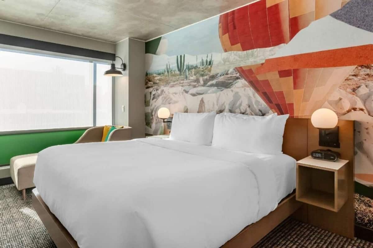 A hotel room with a bed, two bedside lamps, a nightstand, a window, and a mural on the wall.
