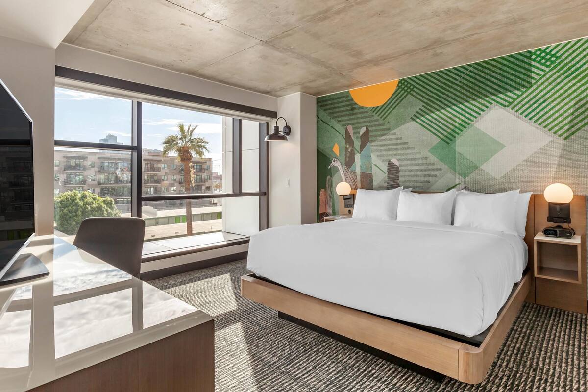 A modern hotel room with a large bed, a desk, and a view of the city through large windows.