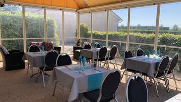 A covered outdoor dining area with tables and chairs, large windows, and a view of greenery.