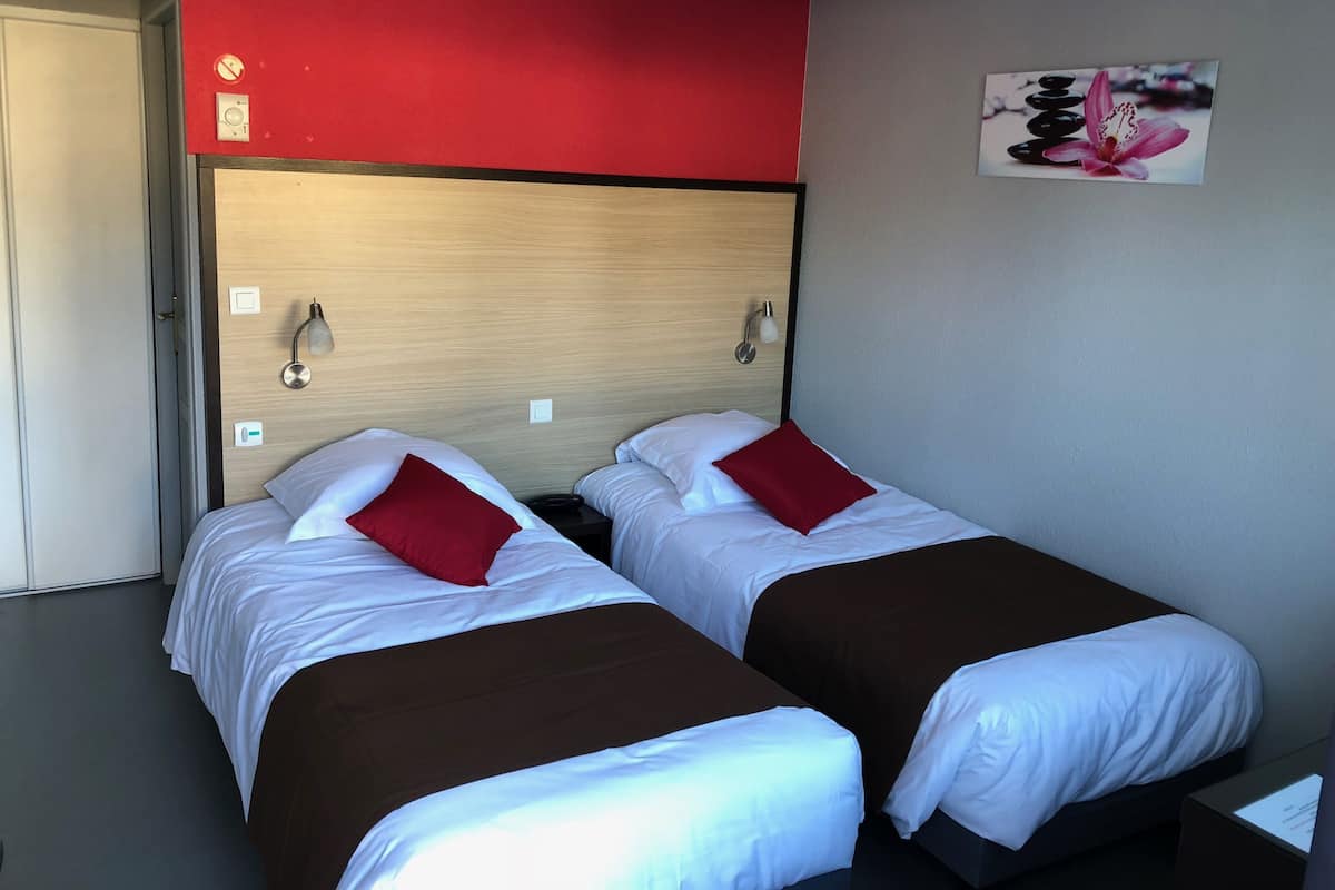 Habitación de hotel con dos camas, cada una con ropa blanca y almohadas rojas, un cabecero de madera y una lámpara fijada a la pared.